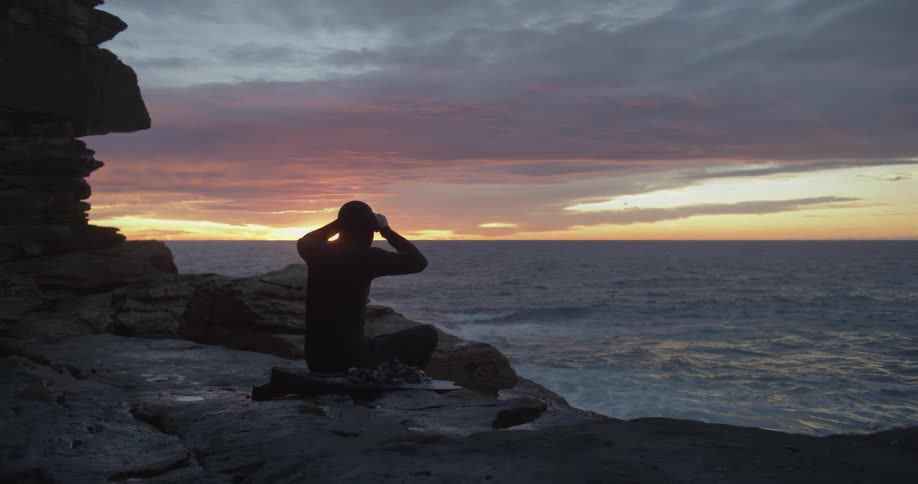 A silhouette of a person with a wetsuit on a rocky coast with the ocean sky and clouds in the background