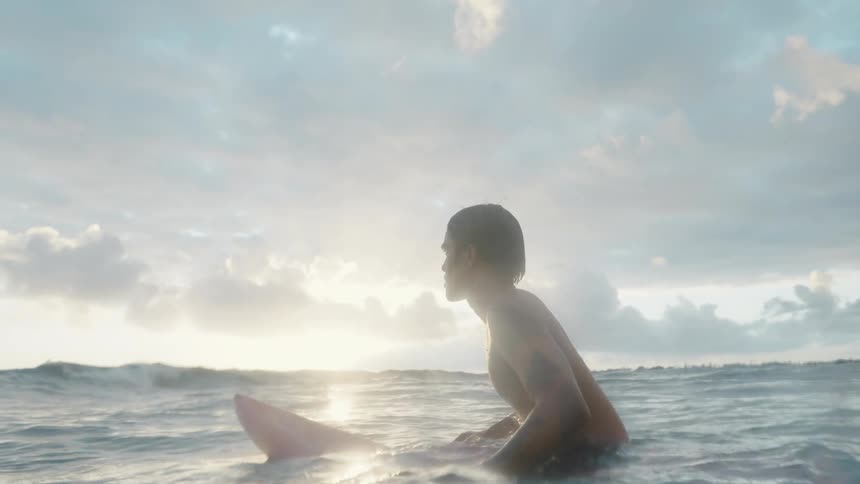 Young tattooed man sitting on a surfboard floating in the ocean at sunset