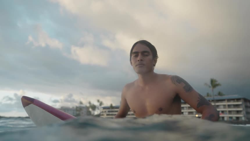 Young tattoed man sitting on a surfboard on the ocean at sunset