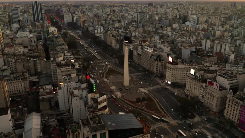 Traffic passes by a tall spire shaped monument in the middle of a dense city