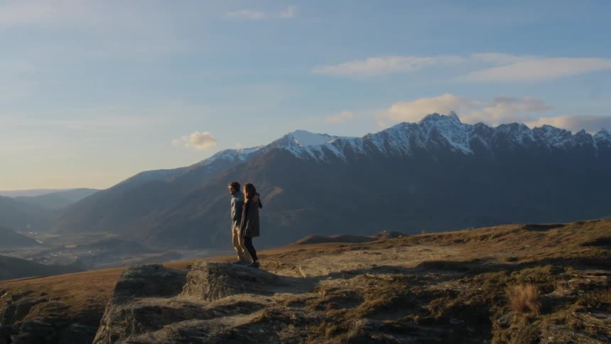 A man and a woman walking and standing on a cliff with a mountain range the sky and clouds in the background