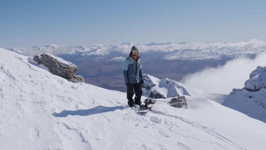 A snowboarder stands at the top of a tall mountain and takes in the view