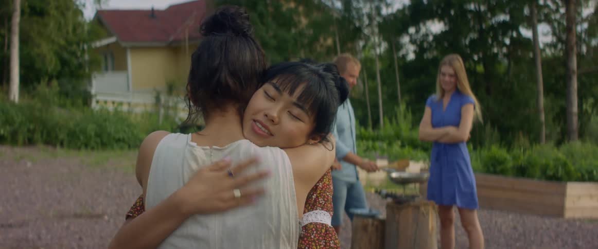 Two women hug as their friends cook outside behind them