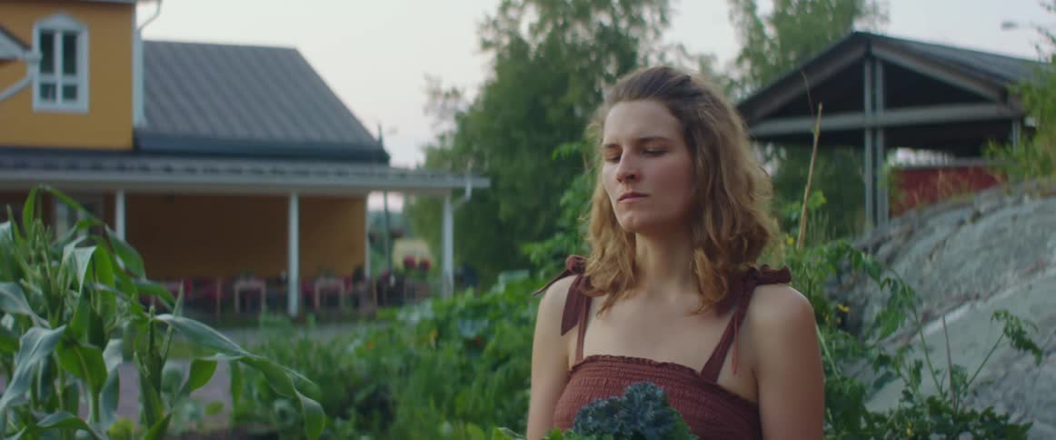 A woman in a garden eating a vegetable with trees the sky and clouds in the background