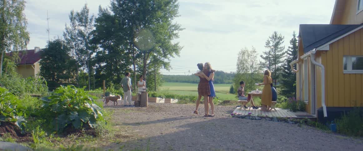 A group of people are standing outside a house or sitting at a table while two women hug one another