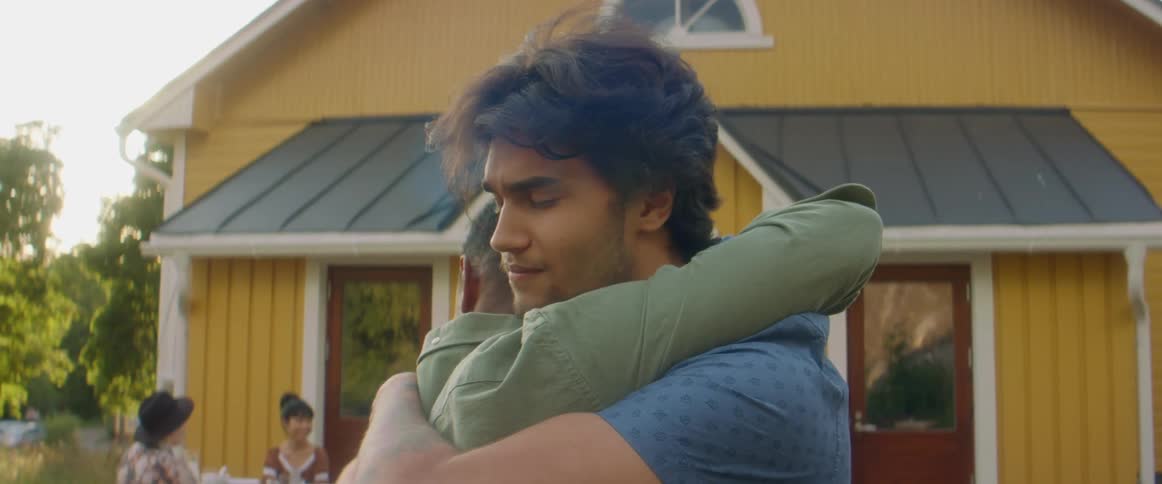 Two men talking and hugging with people sitting at a picnic table in the background