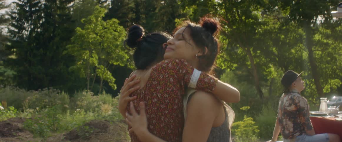 Friends hug standing in yard between set picnic table and small raised plant beds with field and forest in background