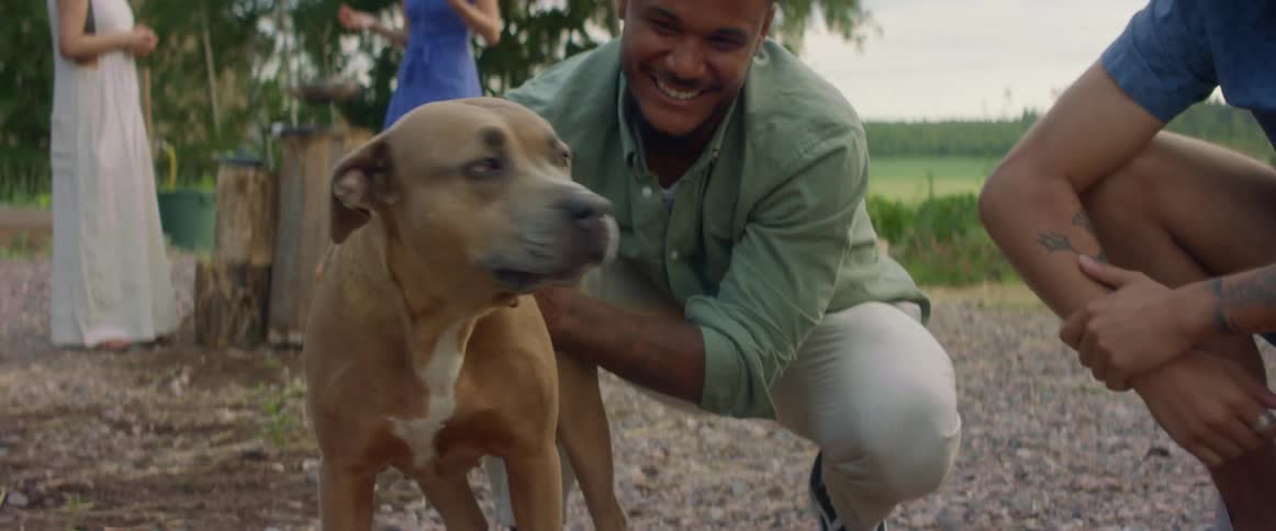 Two women stand and talk as two males crouch petting dog with fenced pasture in the background