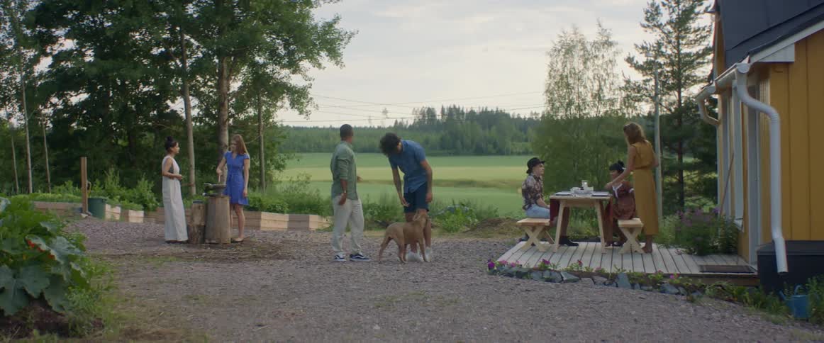 Friends greet each other and pet dog while some of the others sit at picnic table or grill food