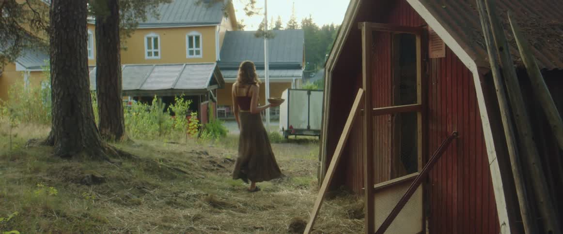 A woman with a basket walking behind a barn with a house trees the sky and clouds in the background