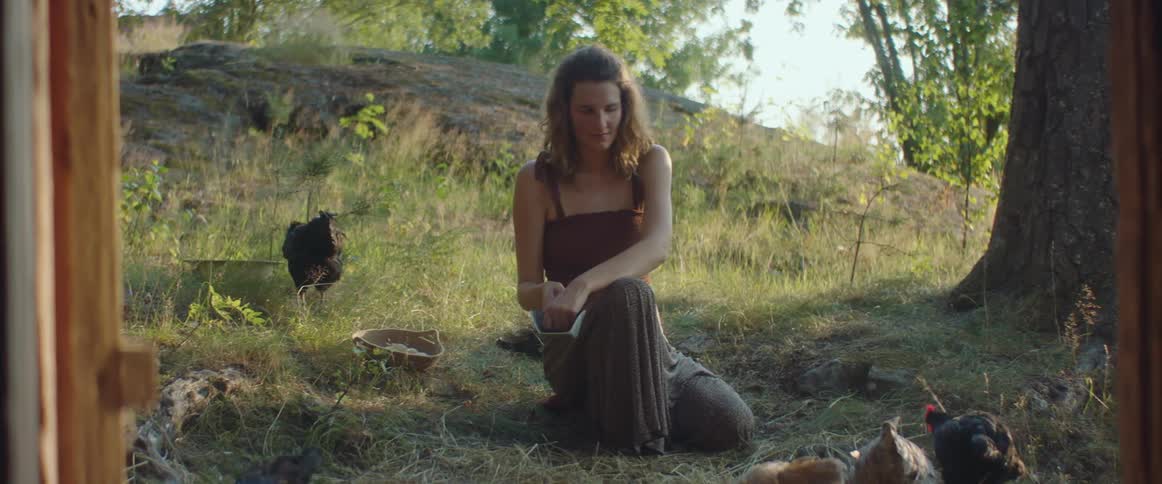 A woman kneels on the ground and sprinkles food to feed her chickens