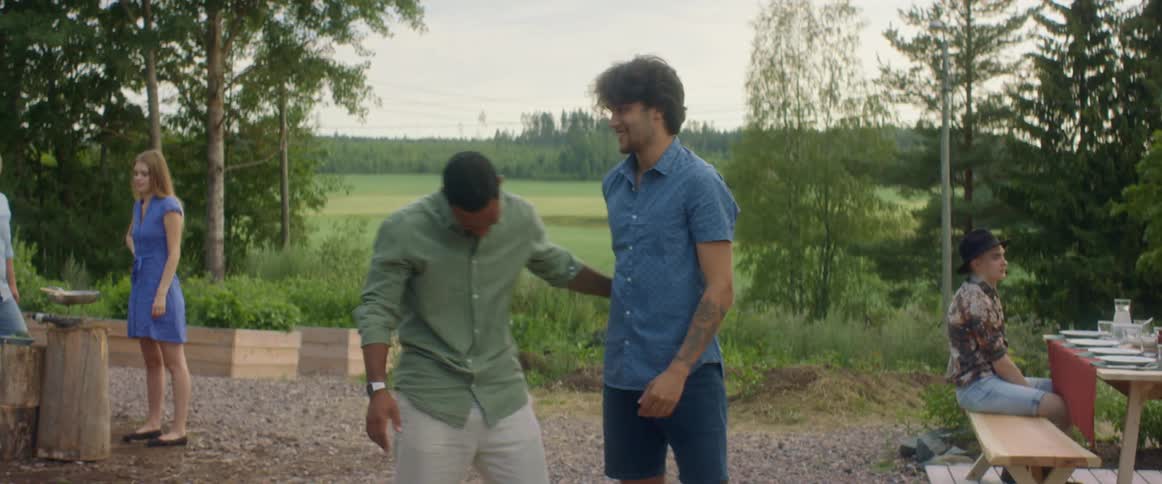 Two men talking and hugging with people trees the sky and clouds in the background