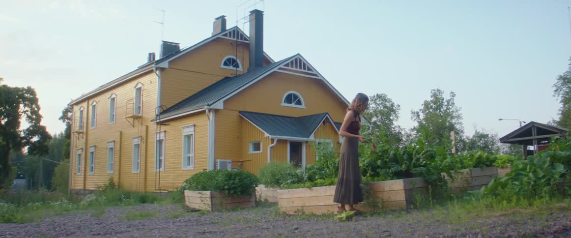 A woman picking vegetables from a garden with a house trees the sky and clouds in the background