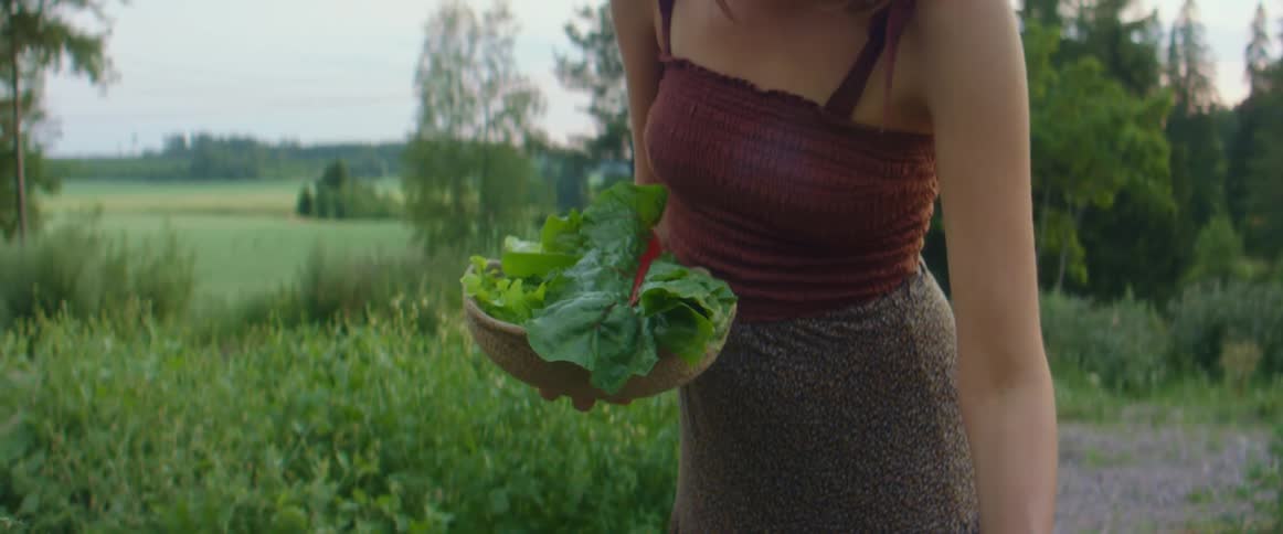A woman in a garden picking vegetables with trees the sky and clouds in the background