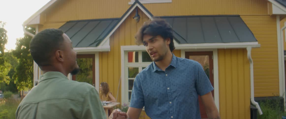 Two male friends shake hands and hug while two women sit at a picnic table behind them