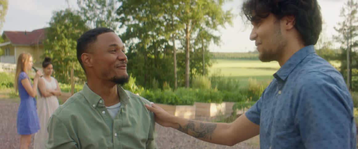 Two men talking and hugging with people trees the sky and clouds in the background
