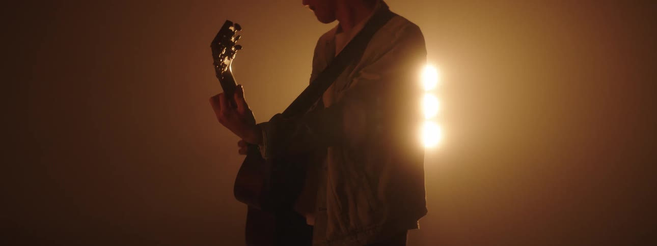 A band member sways as he plays guitar with stage lights glowing behind