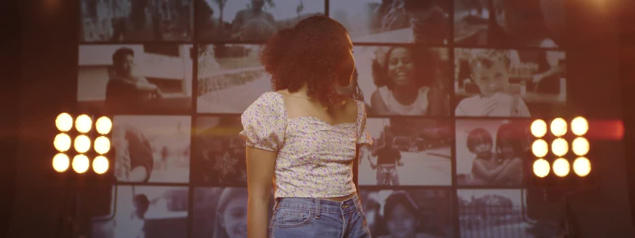 A girl turns her head back from a wall of screens playing video with stage lights glowing