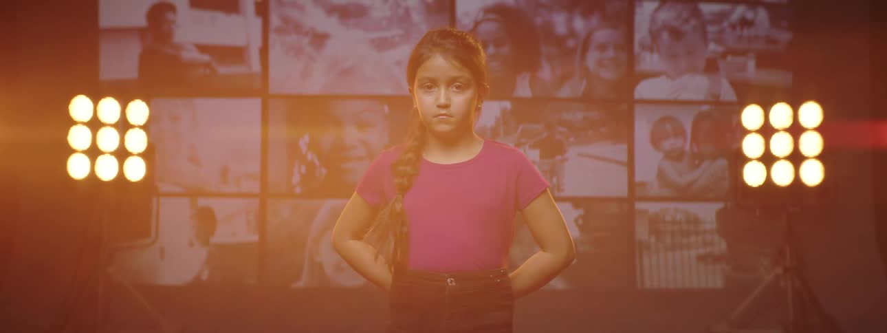 A young girl stands on a stage with electronic screens behind her