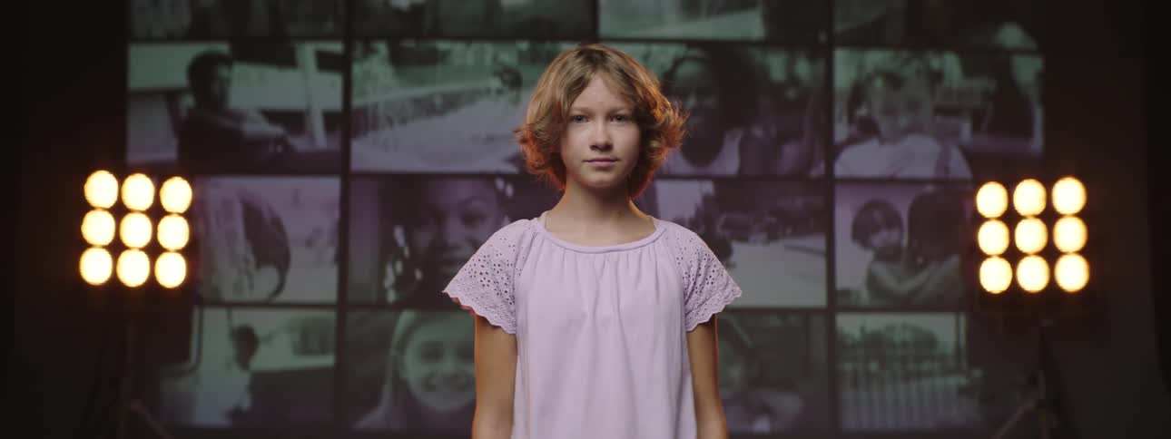 A young woman stares into the camera while standing on a stage with stage lights and a monochrome video wall behind her