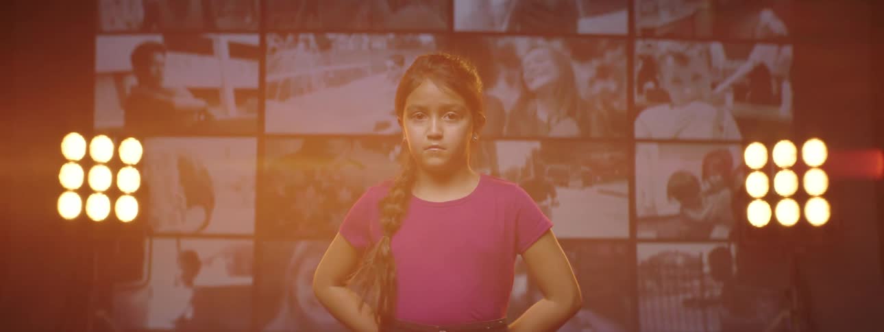 Young girl wearing shorts and a t shirt stands in front of a bank of monitors and some bright lights