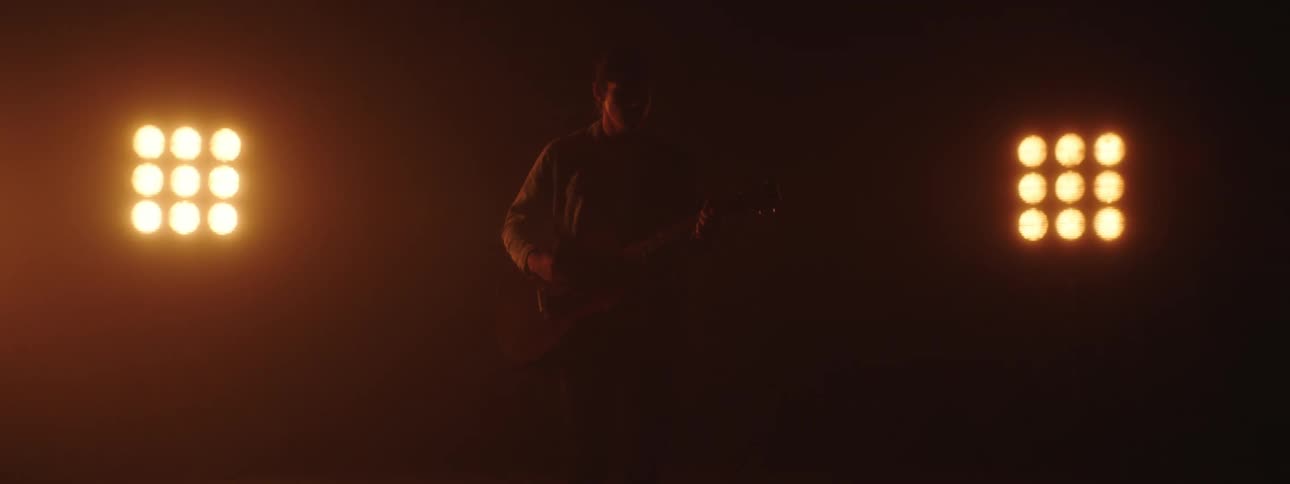 A male guitarist stands and strums his guitar in front of some bright lights