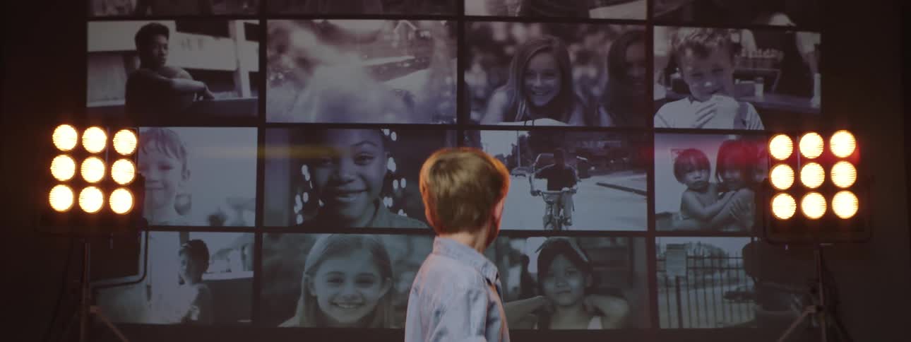 A boy standing before stage lights looks back at a monochrome video wall behind him