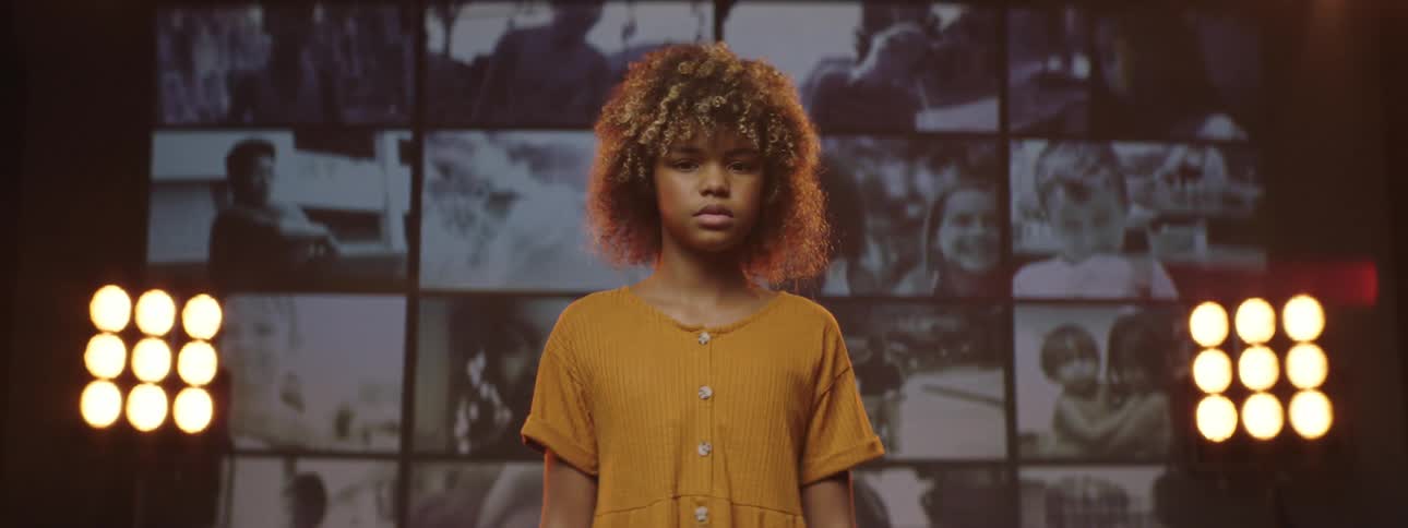 A girl standing and staring on a stage with spotlights a screen and a wall in the background