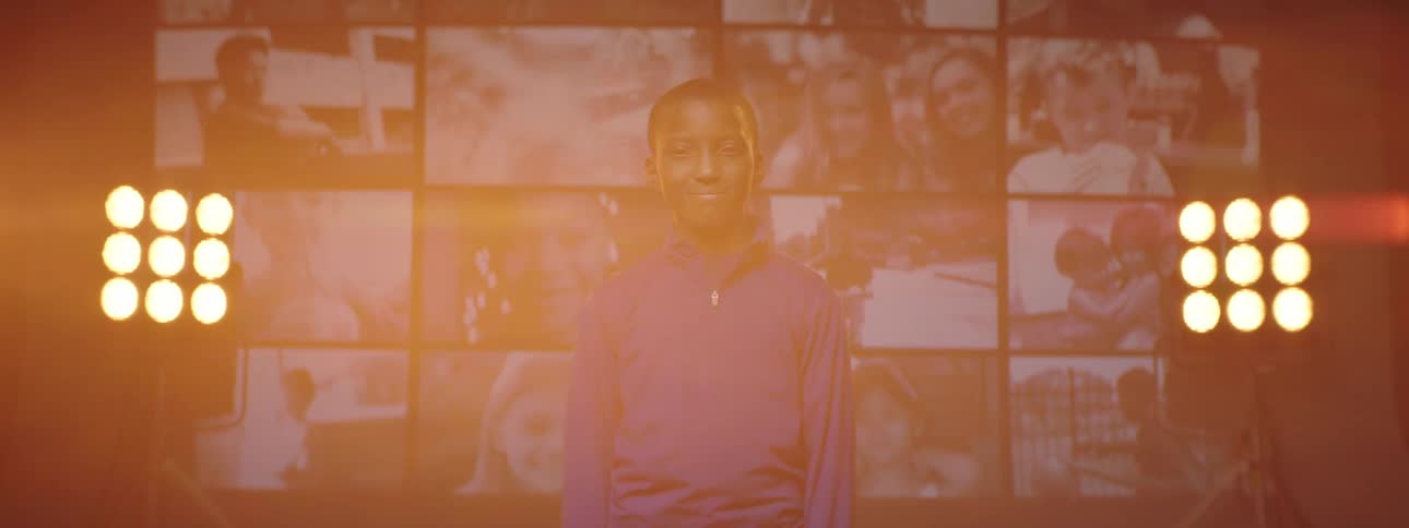 Boy with purple jacket stands facing the camera in front of giant screen with kids