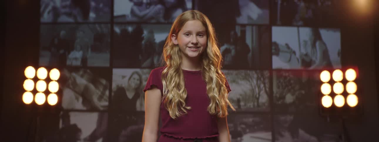A girl standing and staring on a stage with spotlights a screen and a wall in the background
