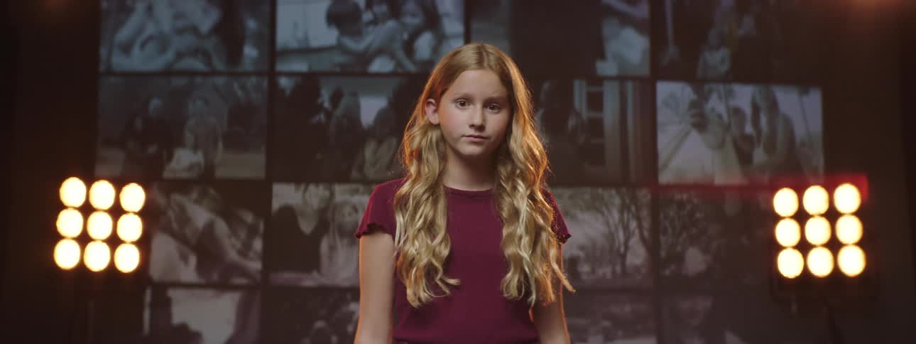 A girl standing and staring on a stage with spotlights a screen and a wall in the background