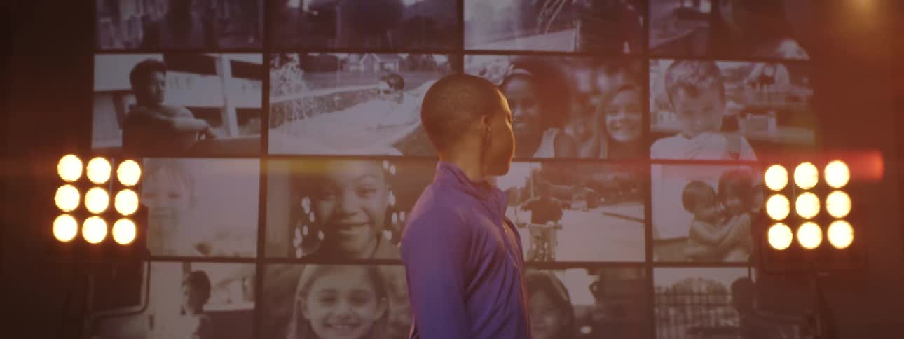 A boy standing and staring on a stage with spotlights a screen and a wall in the background