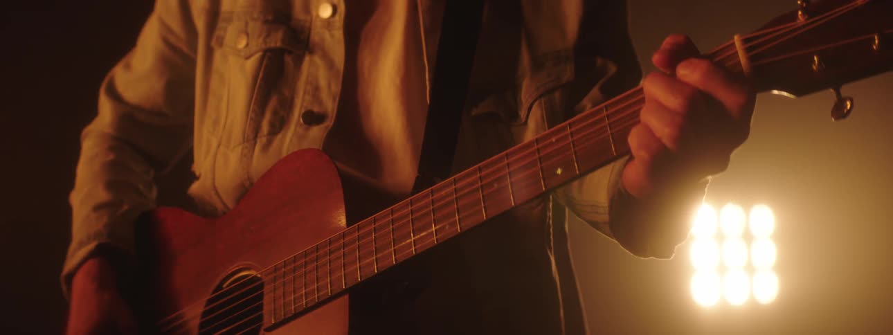 Man wearing a ball cap plays the guitar as bright lights shine in the background