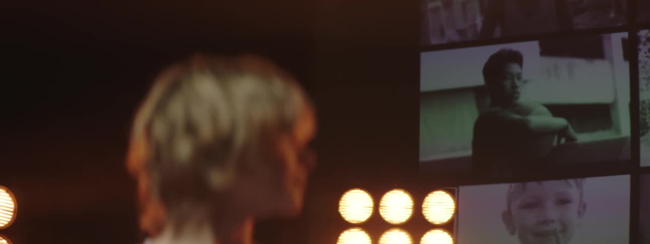 A girl stands with head turned with stage lights and video playing behind her