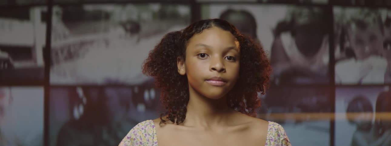Black teenager looks over her shoulder at a wall of monitors then turns her head back to look forward