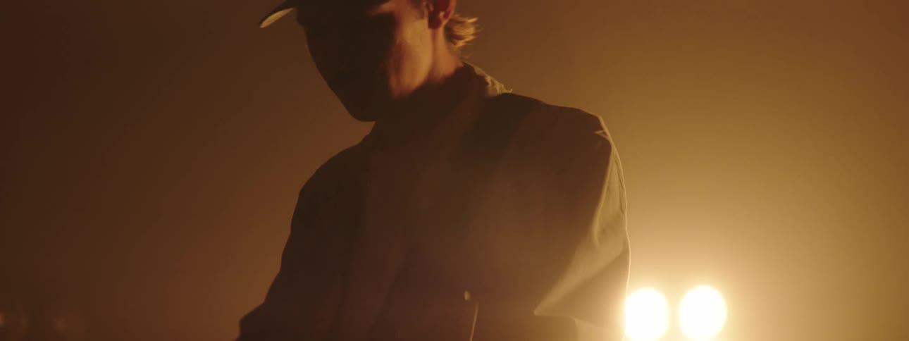 A man with a hat and guitar walking on stage with a spotlight and a wall in the background