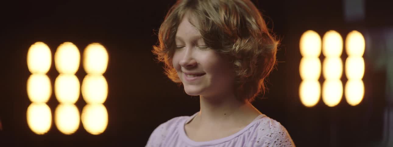 A girl smiles straight ahead with stage lights and video playing behind her