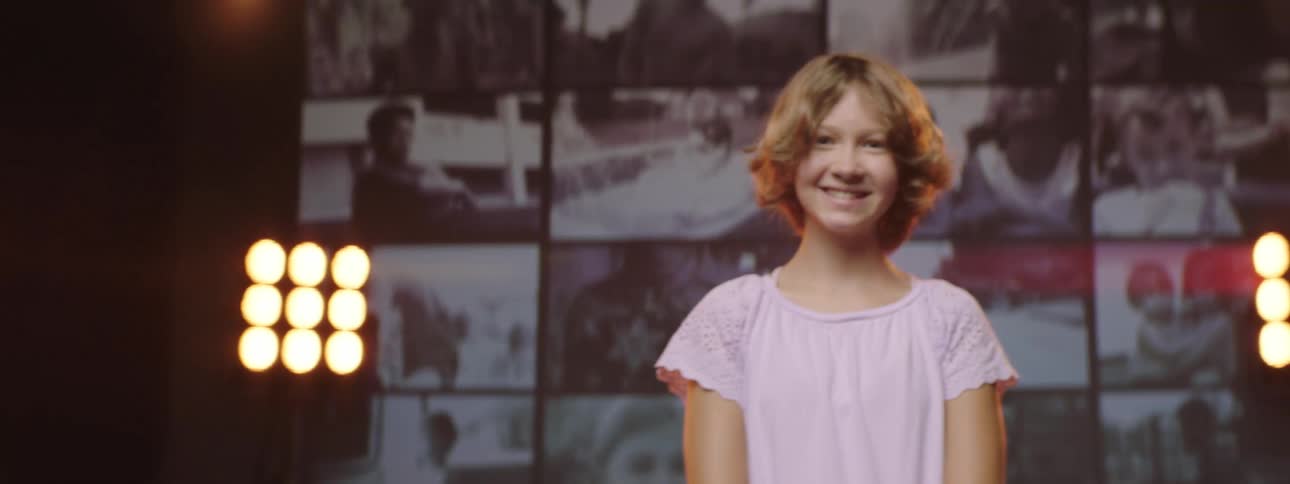A girl standing in front of a wall of video displays smiles