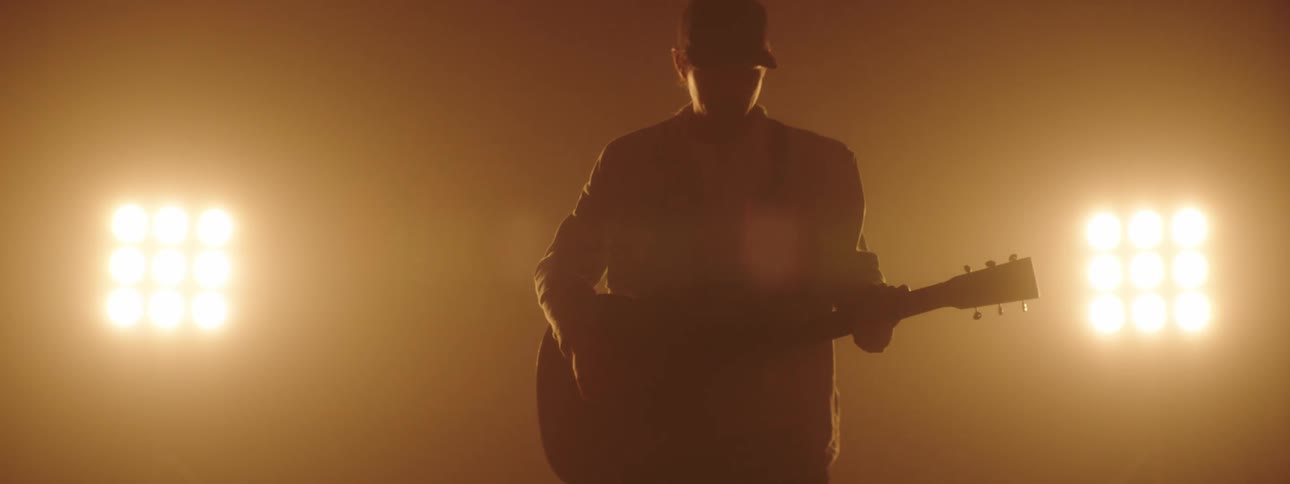 A person with a guitar and a hat on a stage with spotlights and a wall in the background