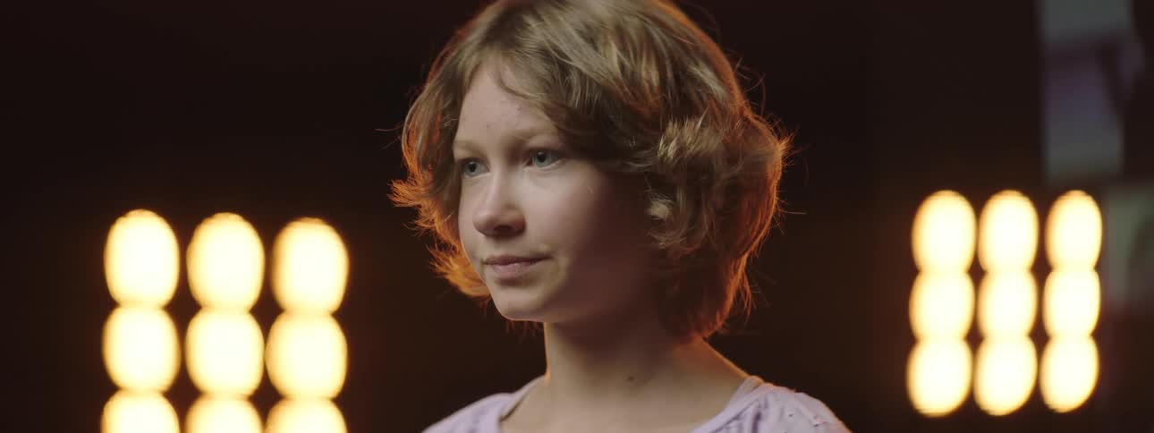 A girl stands motionless before stage lights and a video wall on stage