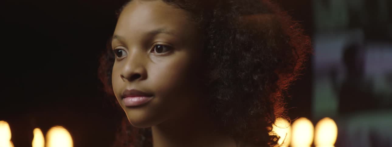 A girl lifts her bowed head and looks straight ahead with video playing behind her