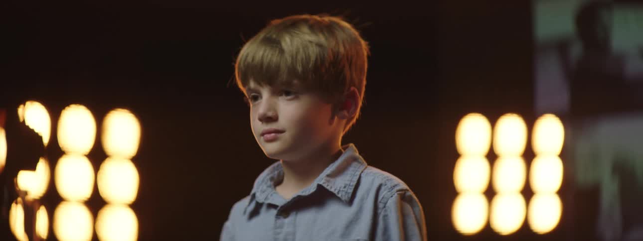 Boy stands on stage backed by stage lights smiling then frowning