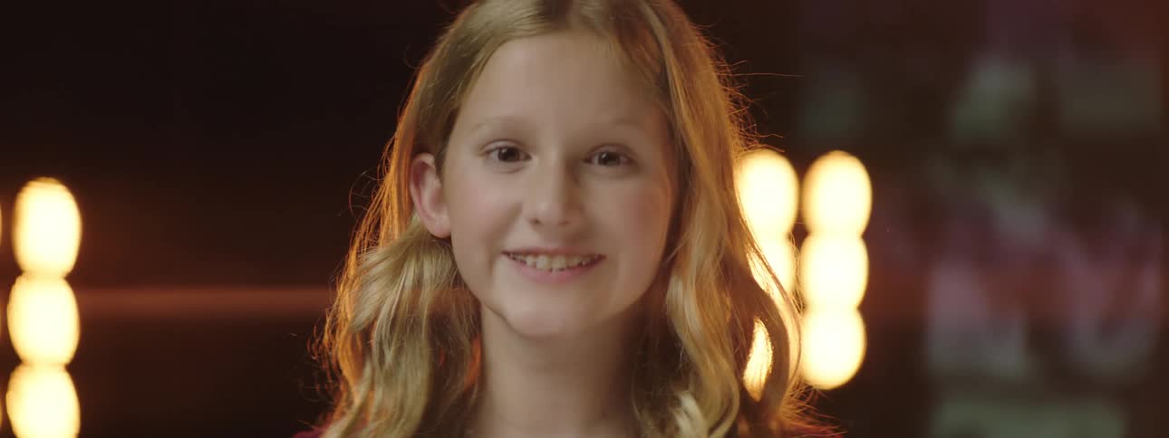 Blonde girl standing on stage looks over shoulder toward digital screens showing videos of people before facing forward and smiling