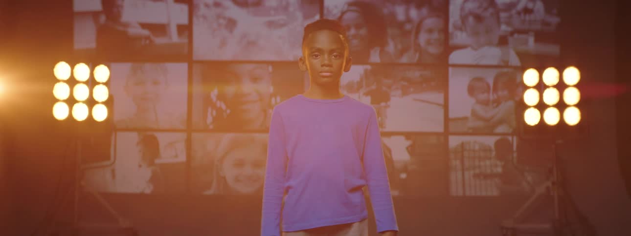Boy stands on stage backed by stage lights and panel of screens showing videos of children