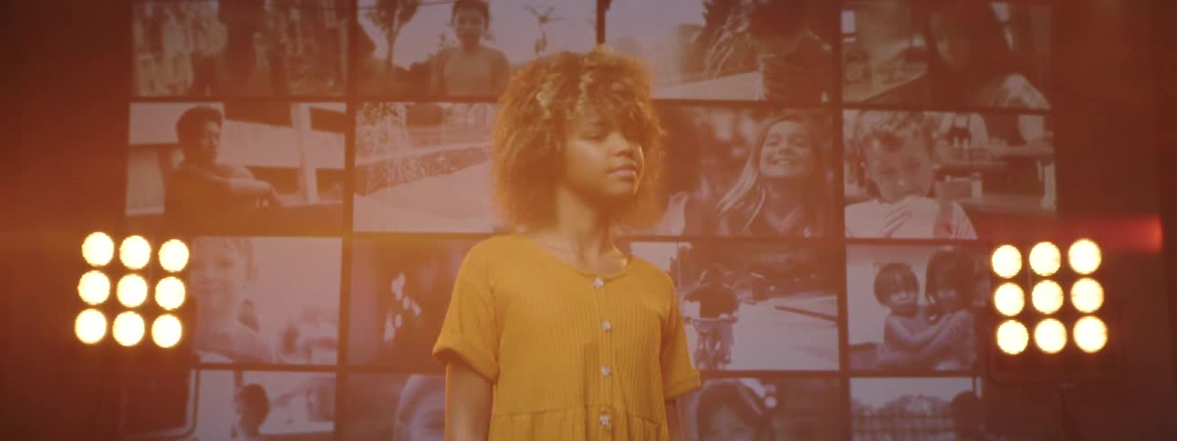 A girl standing and staring on a stage with spotlights a screen and a wall in the background