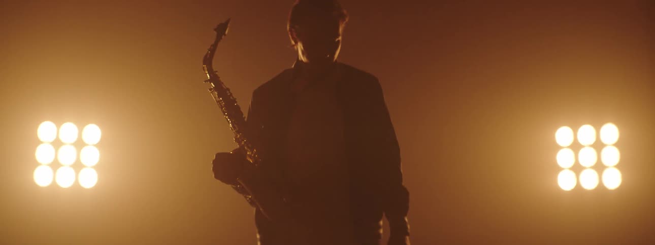 A saxophonist carrying his instrument emerges from the darkness and approaches the camera on stage