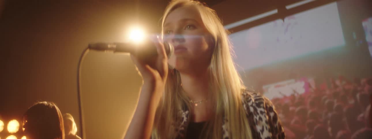 Blonde singer sings smiling as musicians play on stage backed by large wall sized digital screen