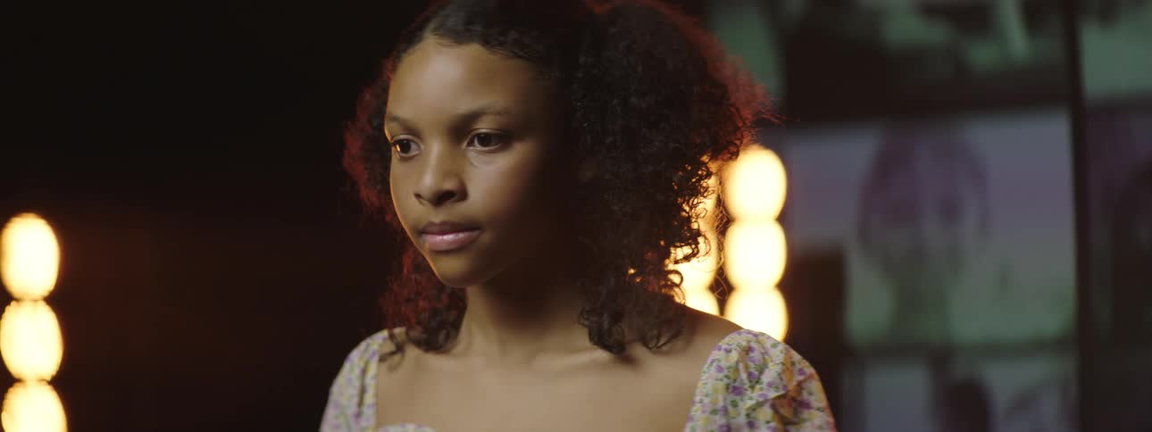 A young woman stands before large stage lights and a video wall in a large dark room