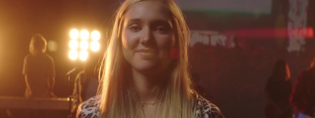Blonde female singer smiles holding out microphone on stage backed by digital screen as rest of band stands