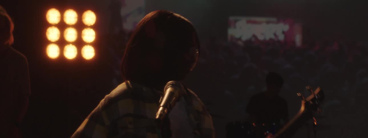 A female plays guitar on a stage with other musicians with bright lights behind them